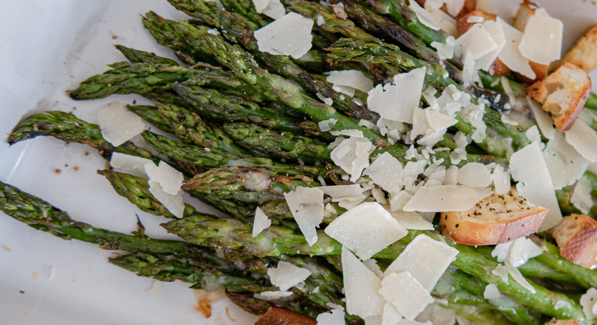 Roasted asparagus spears topped with shaved parmesan cheese and pieces of toasted croutons on a white plate.
