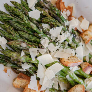 Roasted asparagus spears topped with shaved Parmesan cheese and croutons, served in a white dish.