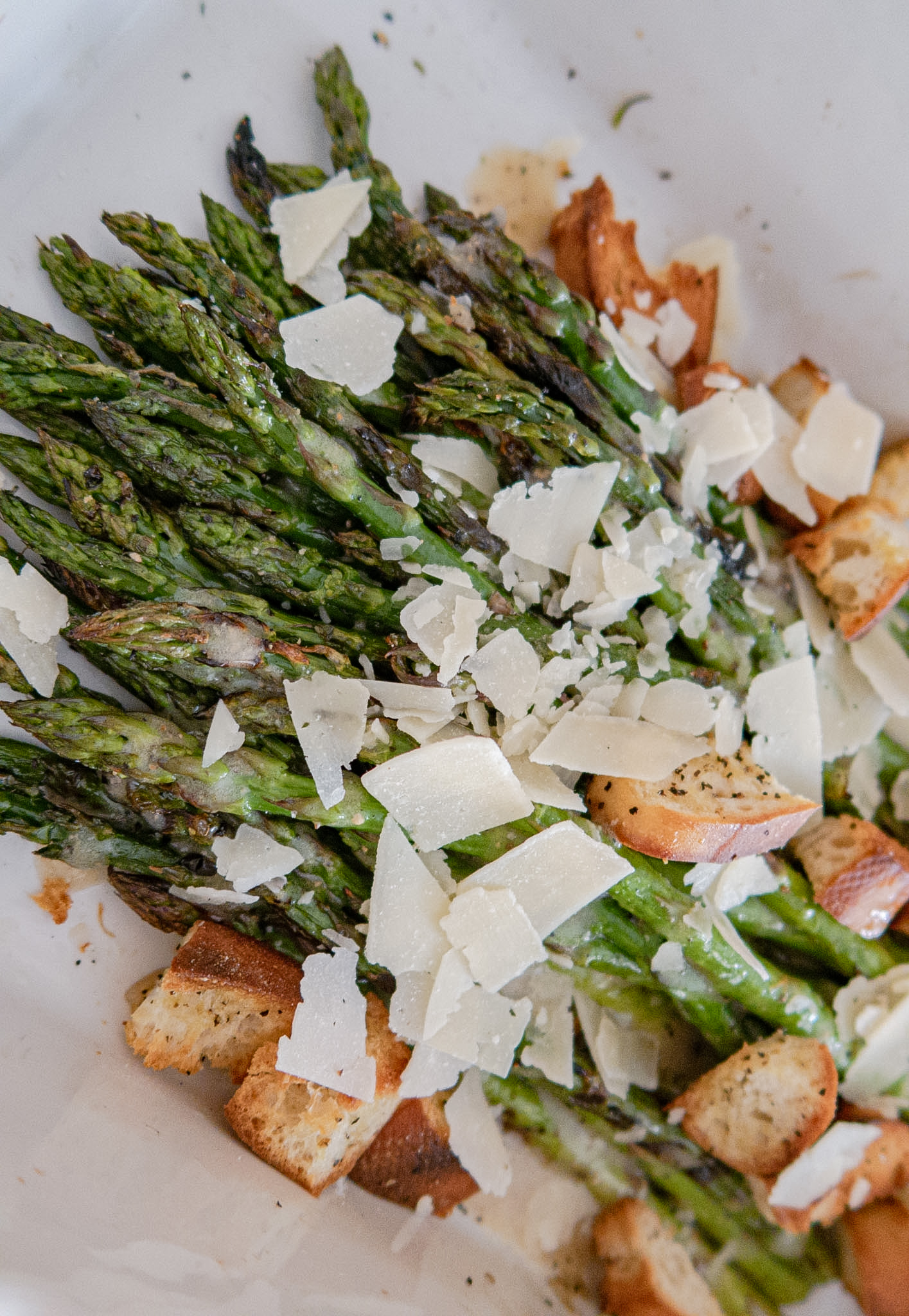 Roasted asparagus spears topped with shaved Parmesan cheese and croutons, served in a white dish.