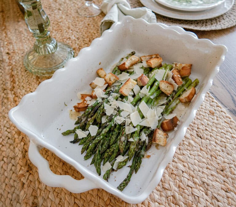 A white ceramic baking dish filled with roasted asparagus, topped with shaved parmesan cheese and croutons, sits on a woven placemat on a dining table.