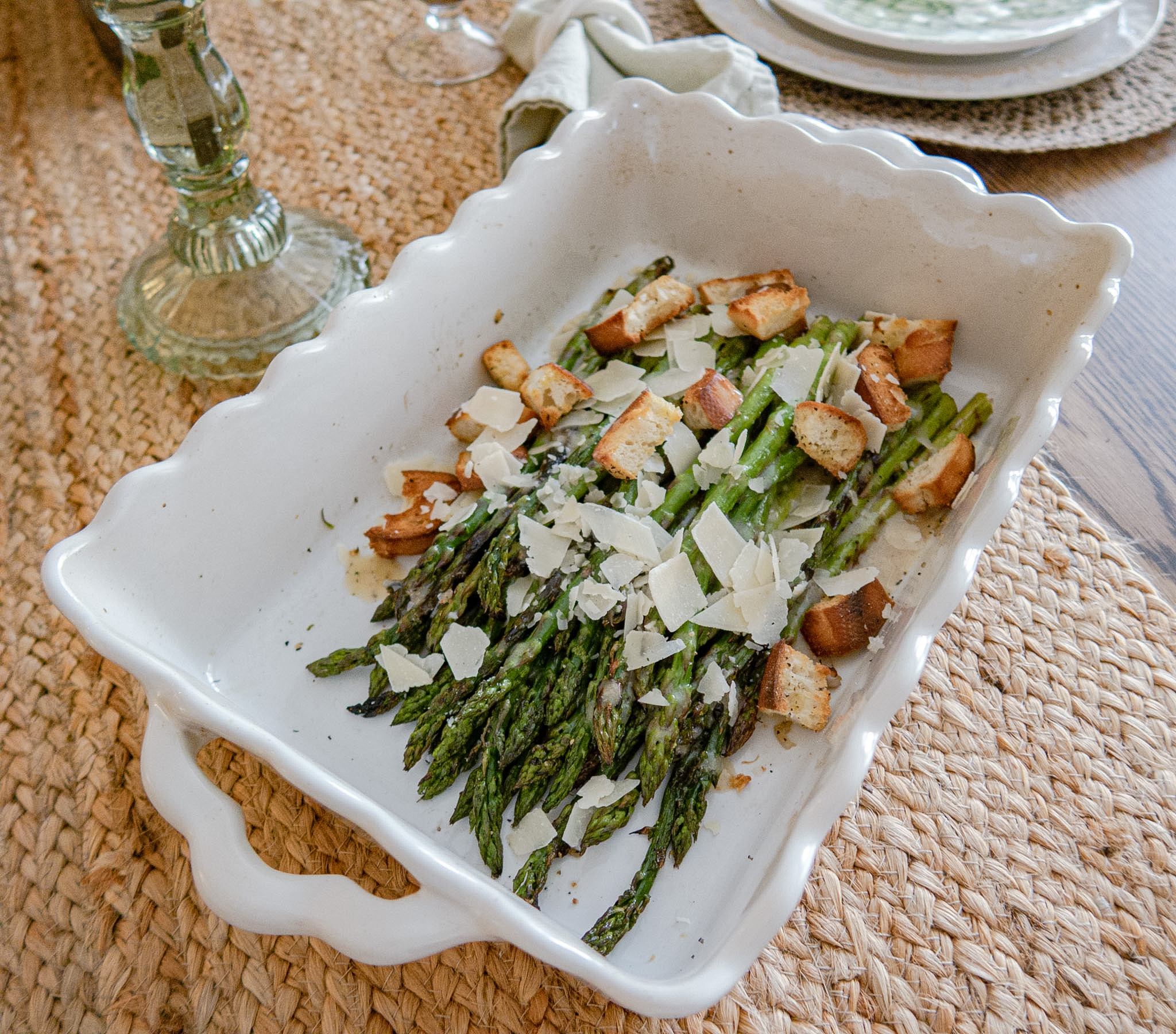 A white ceramic baking dish filled with roasted asparagus, topped with shaved parmesan cheese and croutons, sits on a woven placemat on a dining table.