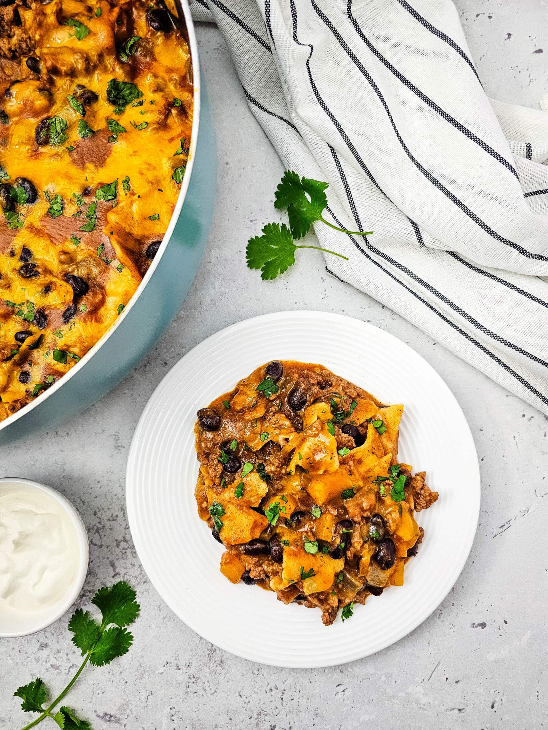 A white plate with a serving of cheesy baked casserole with black beans and herbs, next to a baking dish, a striped towel, fresh cilantro, and a small bowl of sour cream.