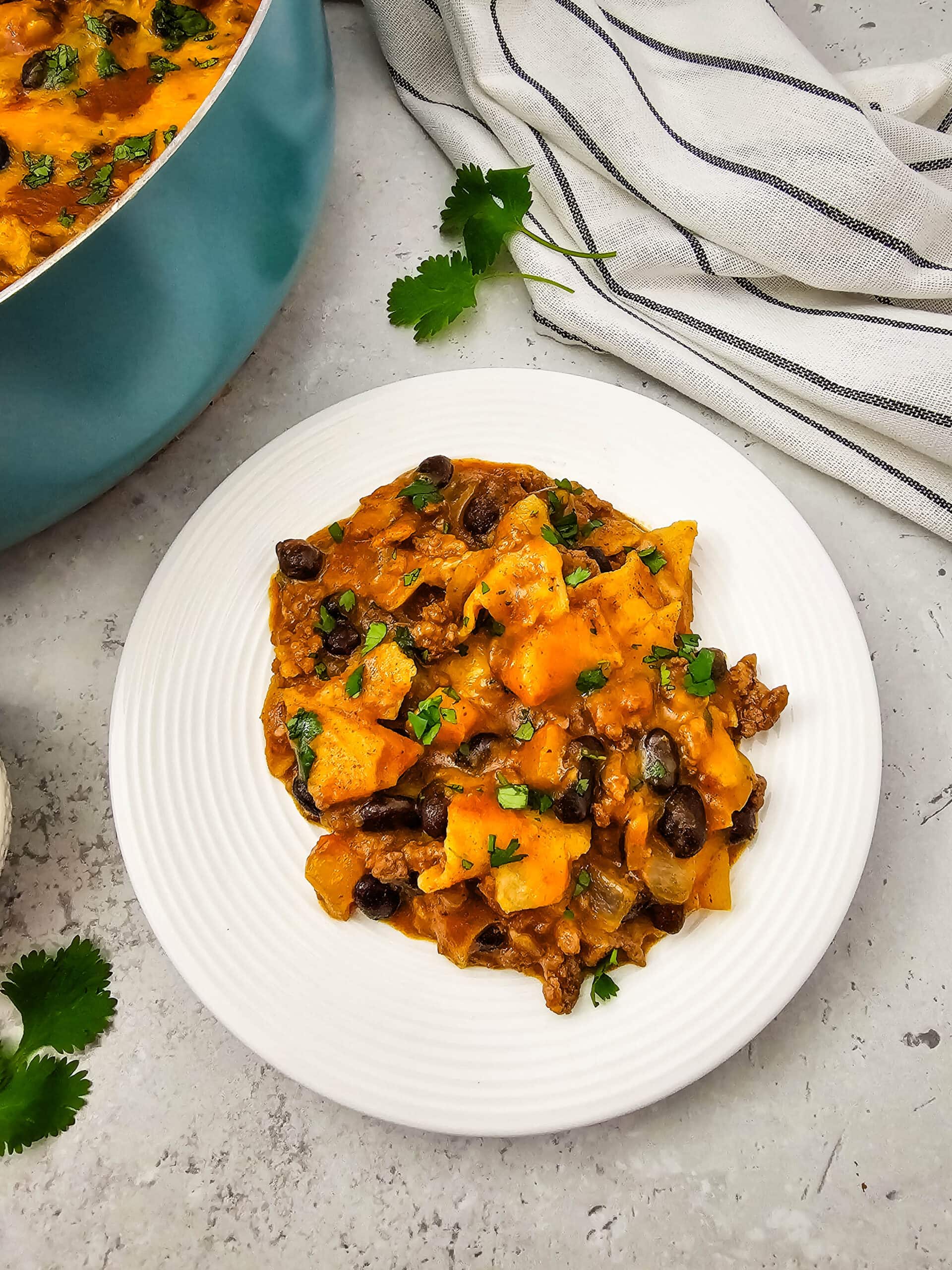 A white plate with a serving of cheesy tortilla casserole with black beans, ground meat, and chopped cilantro. A pot with more casserole and a striped towel are in the background.