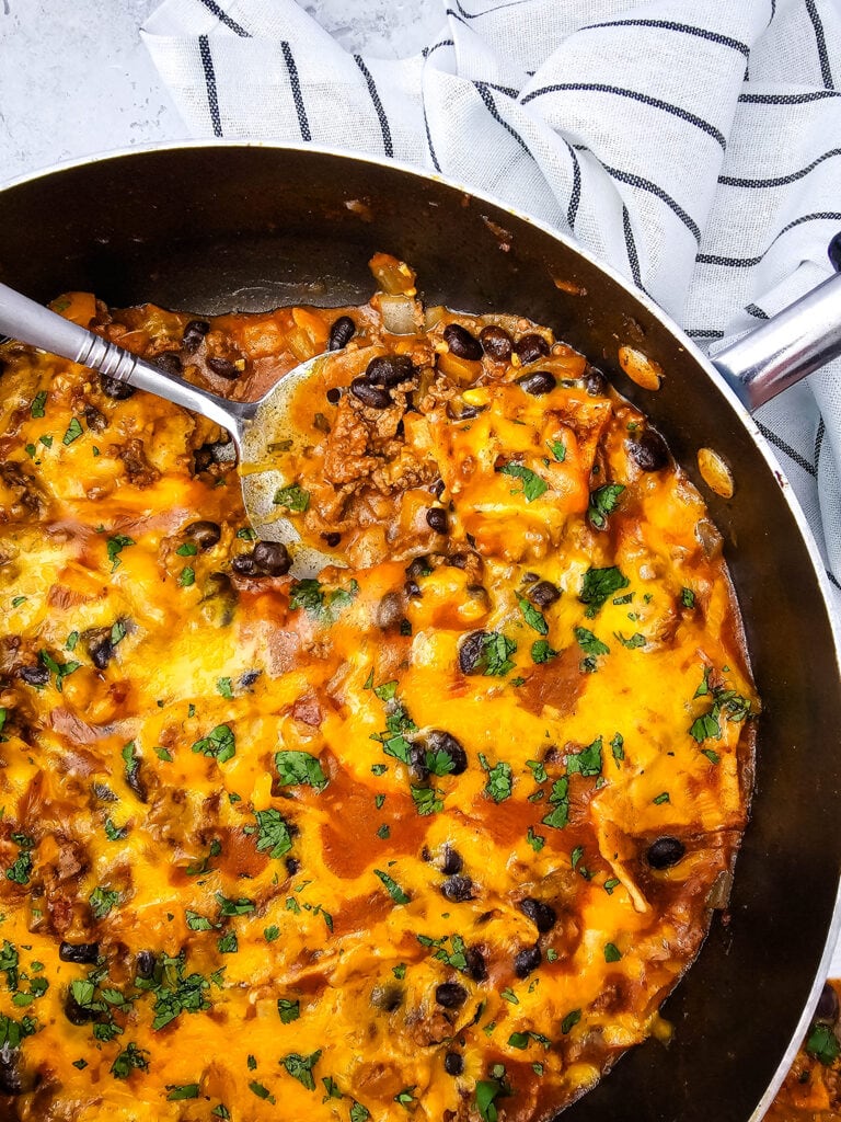 A skillet filled with cheesy beef, black bean, and tortilla casserole, topped with melted cheese and chopped herbs. A spoon rests inside, and a striped towel is nearby.