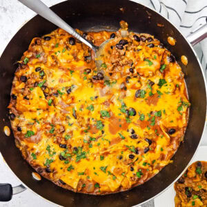 A skillet filled with cheesy taco casserole, topped with melted cheese and fresh herbs, with a serving spoon and a portion scooped onto a plate nearby.