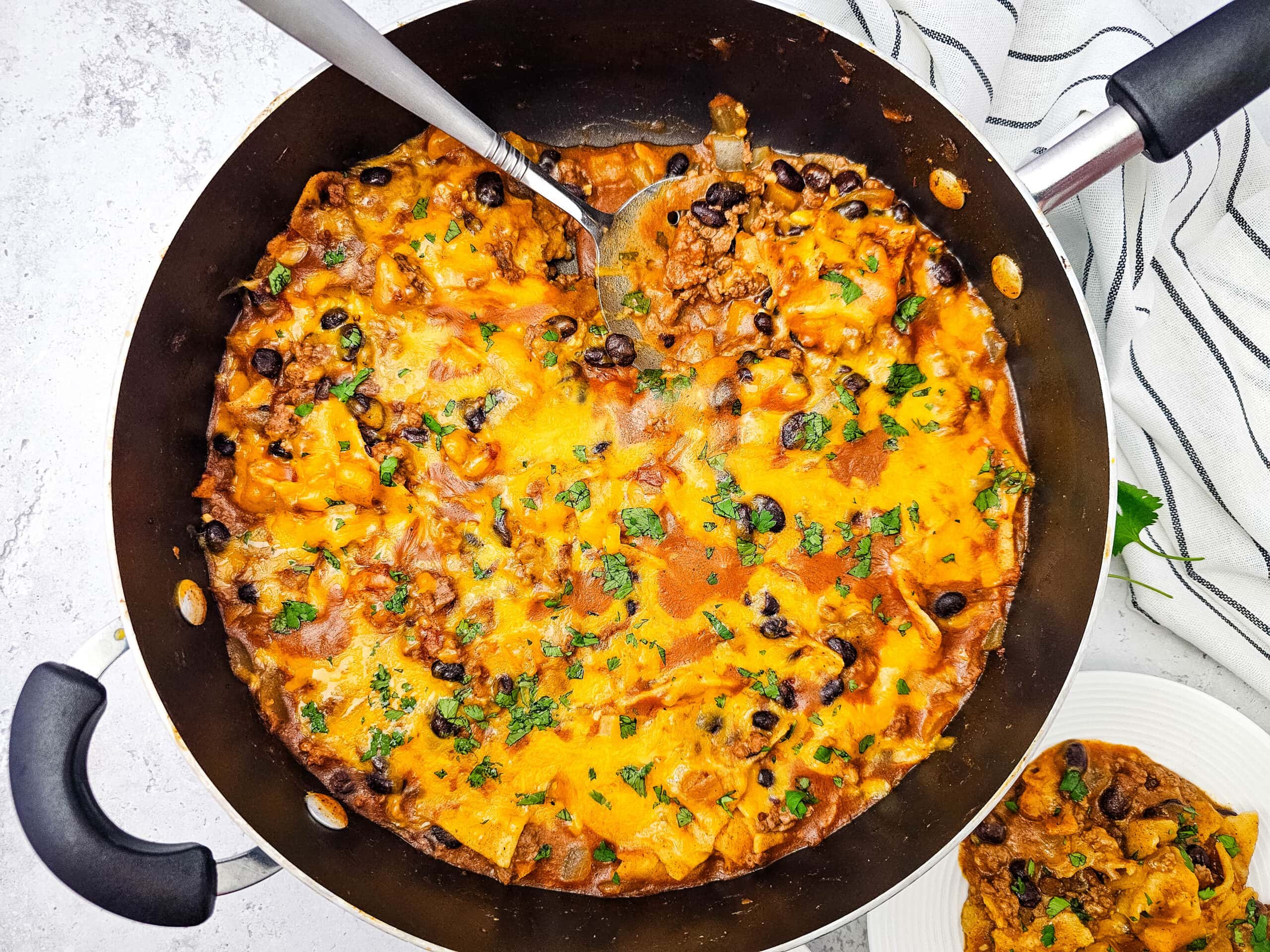 A skillet filled with cheesy taco casserole, topped with melted cheese and fresh herbs, with a serving spoon and a portion scooped onto a plate nearby.