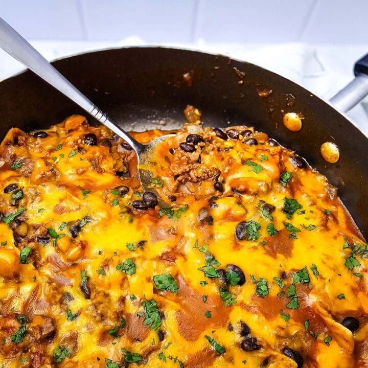 A skillet filled with cheesy beef and black bean casserole, topped with melted cheese and chopped herbs. A spatula is lifting a portion, revealing layers of meat, sauce, and beans.