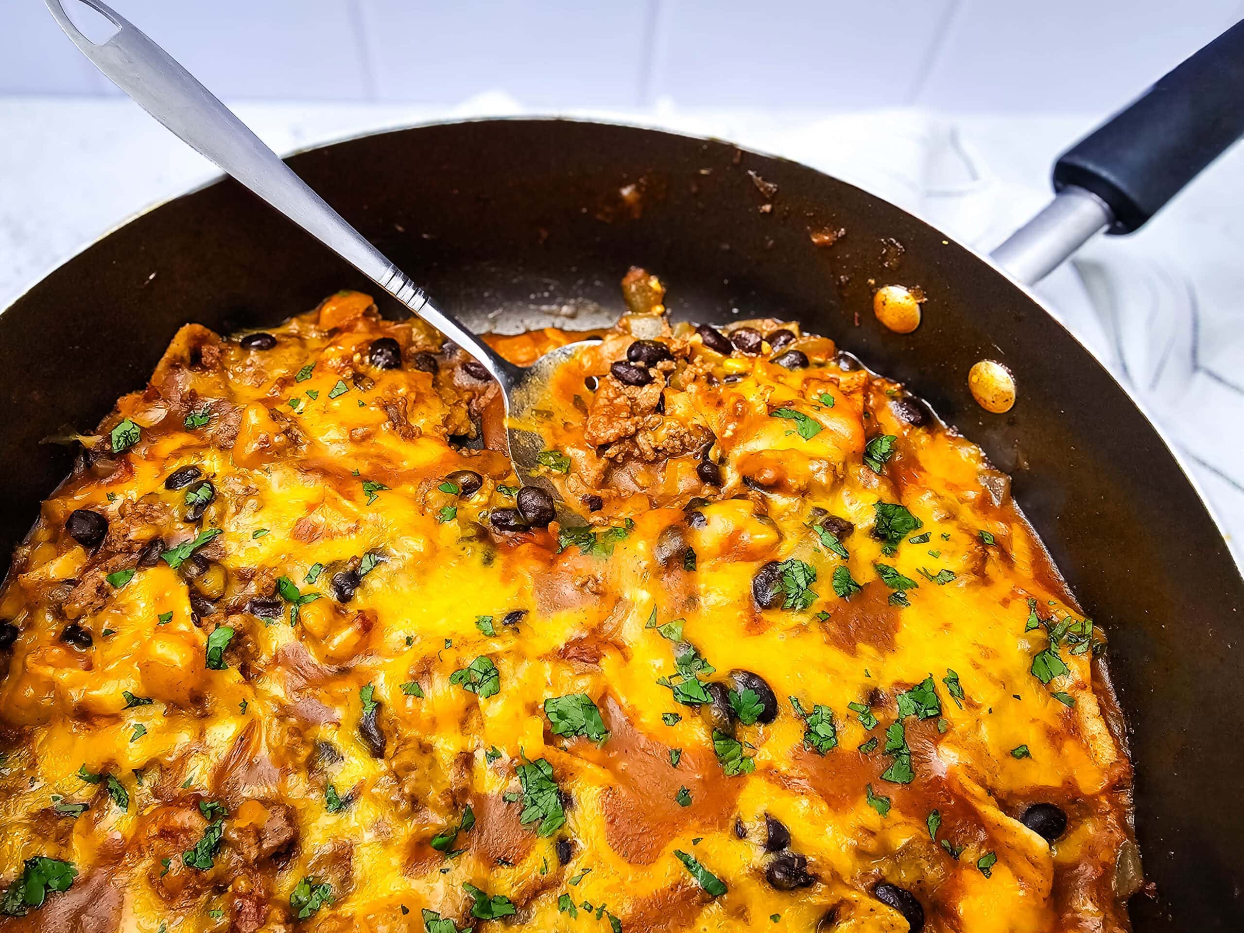 A skillet filled with cheesy beef and black bean casserole, topped with melted cheese and chopped herbs. A spatula is lifting a portion, revealing layers of meat, sauce, and beans.