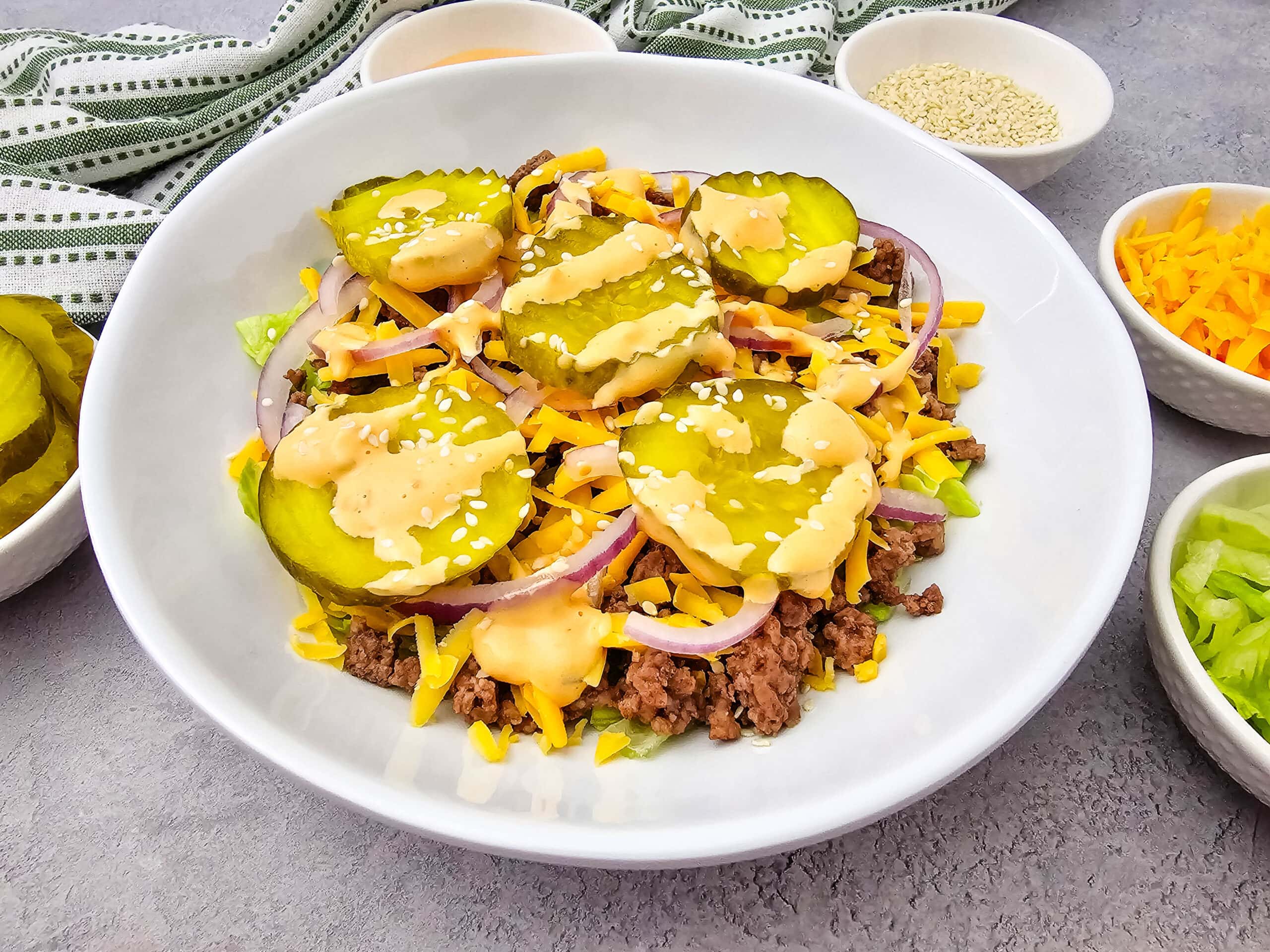 A white bowl filled with ground beef, shredded cheese, lettuce, red onion, pickles, and a drizzle of sauce, topped with sesame seeds. Small bowls of cheese, lettuce, and seeds are nearby.