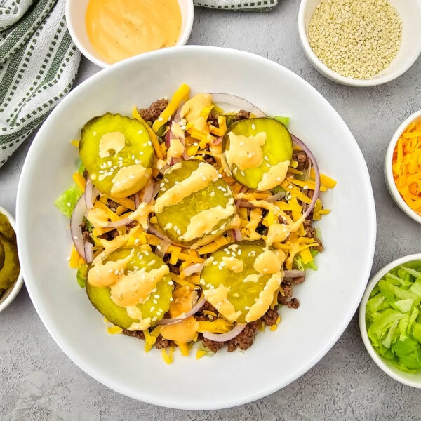 A salad topped with pickles, shredded cheese, onions, sesame seeds, and dressing is surrounded by small bowls of sauce, sesame seeds, cheese, pickles, and lettuce on a gray surface.