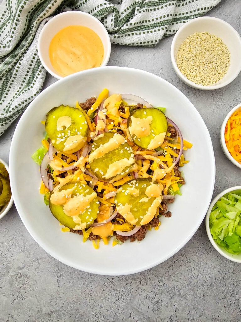 A salad topped with pickles, shredded cheese, onions, sesame seeds, and dressing is surrounded by small bowls of sauce, sesame seeds, cheese, pickles, and lettuce on a gray surface.