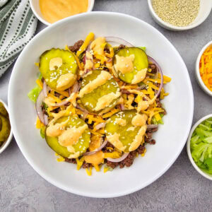 A bowl with ground beef, lettuce, shredded cheese, pickles, onions, sesame seeds, and dressing. Surrounding bowls hold extra pickles, shredded cheese, lettuce, sesame seeds, and dressing. A striped cloth is nearby.