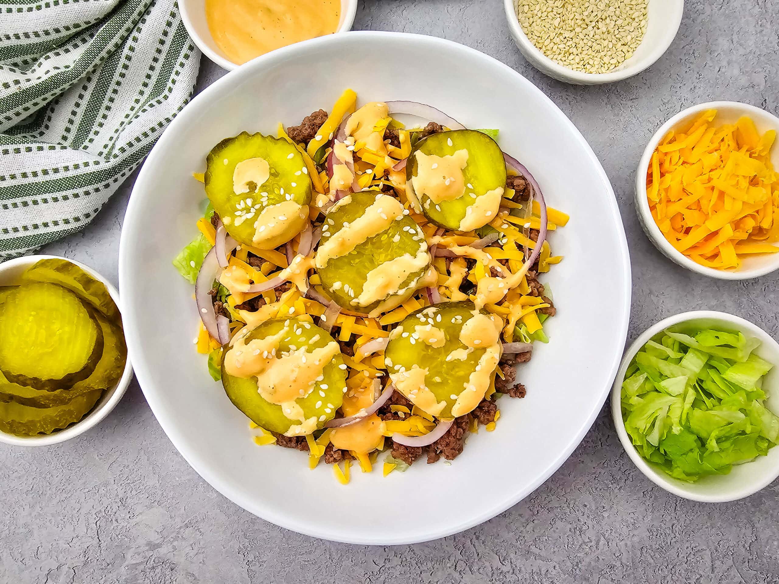 A bowl with ground beef, lettuce, shredded cheese, pickles, onions, sesame seeds, and dressing. Surrounding bowls hold extra pickles, shredded cheese, lettuce, sesame seeds, and dressing. A striped cloth is nearby.