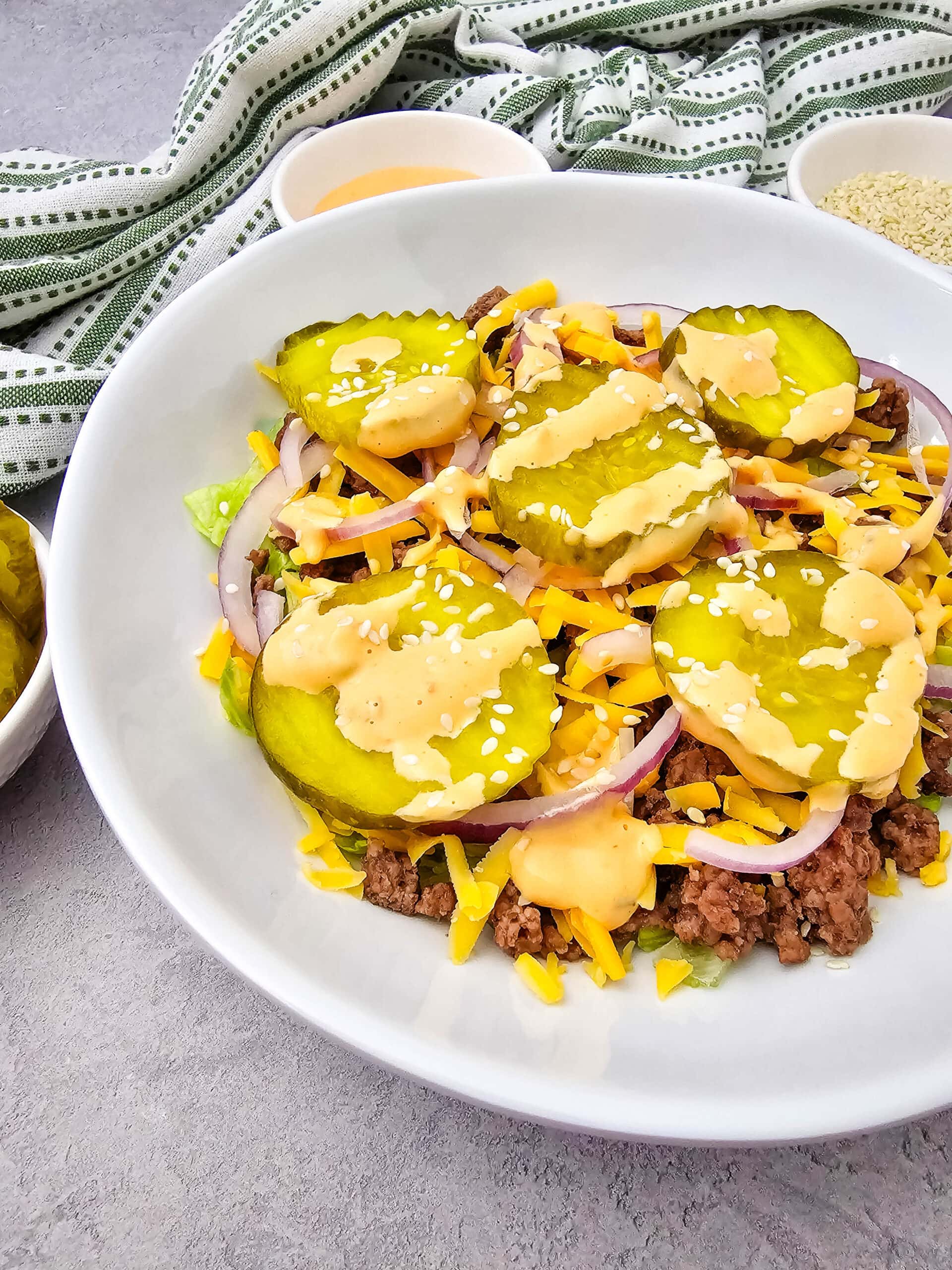 A bowl with ground beef, shredded cheddar cheese, sliced red onions, pickles, sesame seeds, and creamy dressing, arranged on a bed of lettuce. A green-striped towel and condiment dishes are in the background.