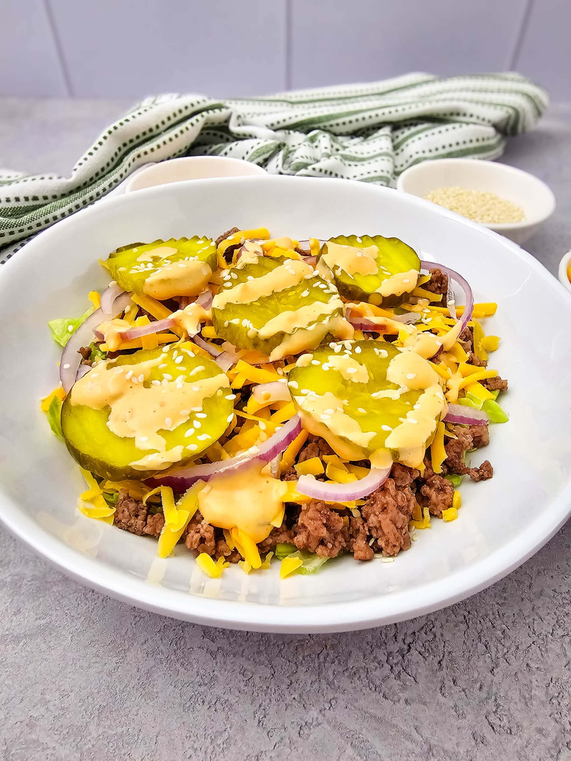 A bowl of ground beef, shredded cheddar cheese, pickles, red onion, lettuce, sesame seeds, and creamy sauce, arranged like a deconstructed cheeseburger.