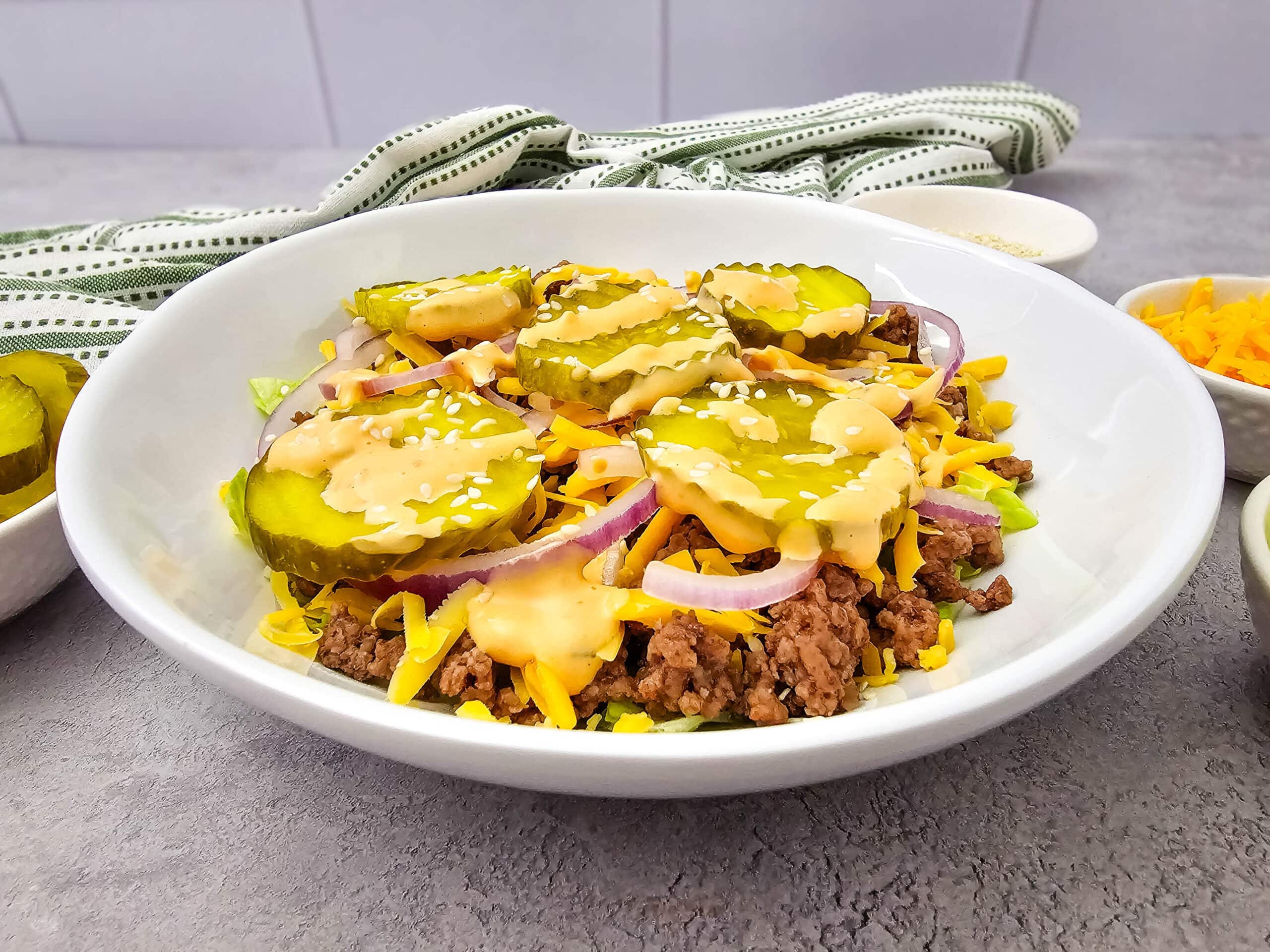 A white bowl filled with ground beef, shredded cheese, sliced red onions, pickles, and sauce, garnished with sesame seeds. A green-striped cloth and small bowls of cheese and pickles are in the background.