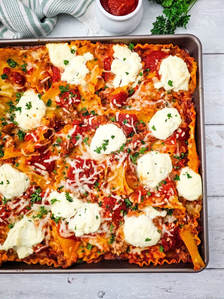 Baking sheet of lasagna topped with melted cheese, tomato sauce, and dollops of ricotta, garnished with chopped parsley, on a white wooden table.