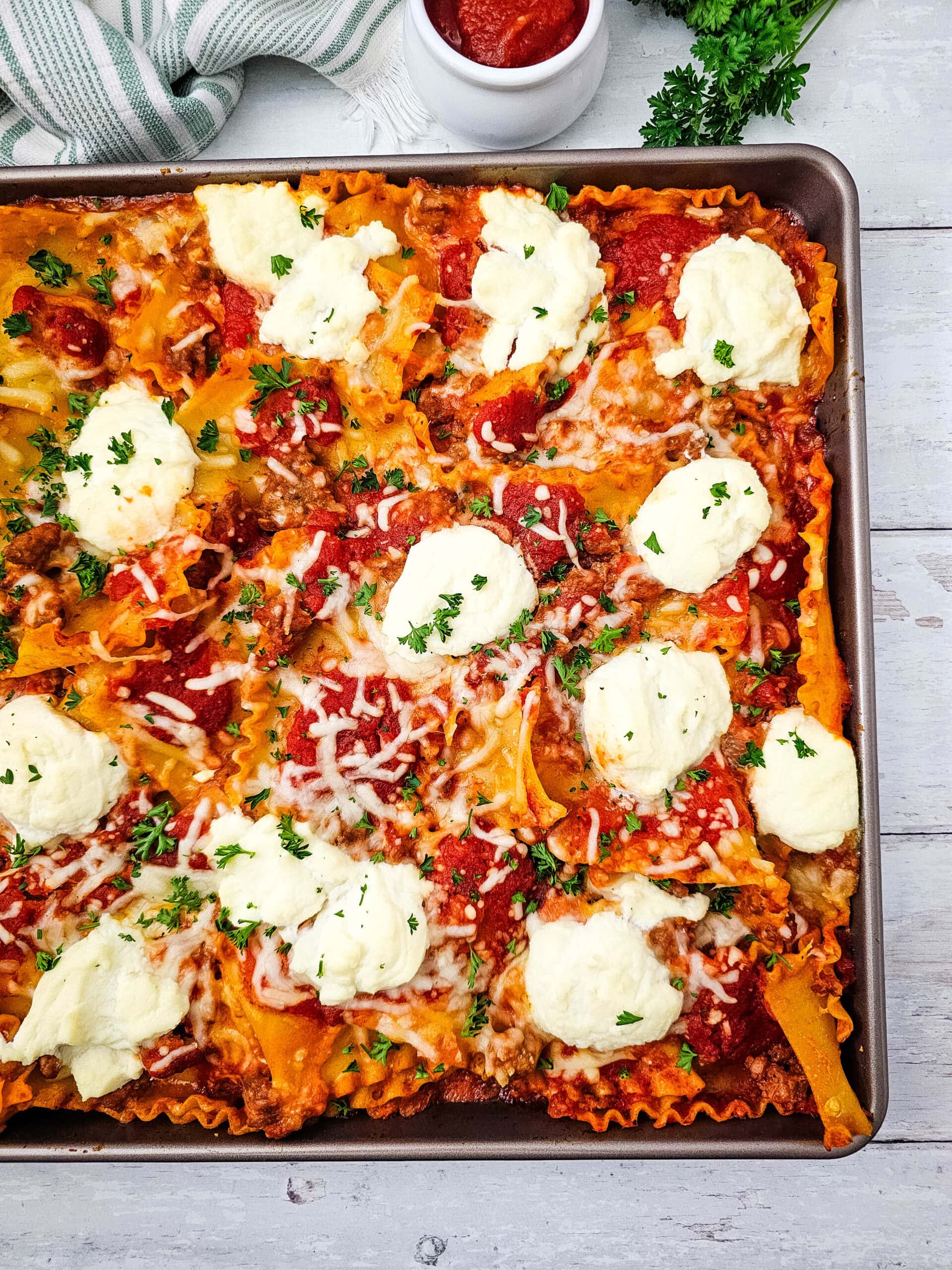 Baking sheet of lasagna topped with melted cheese, tomato sauce, and dollops of ricotta, garnished with chopped parsley, on a white wooden table.