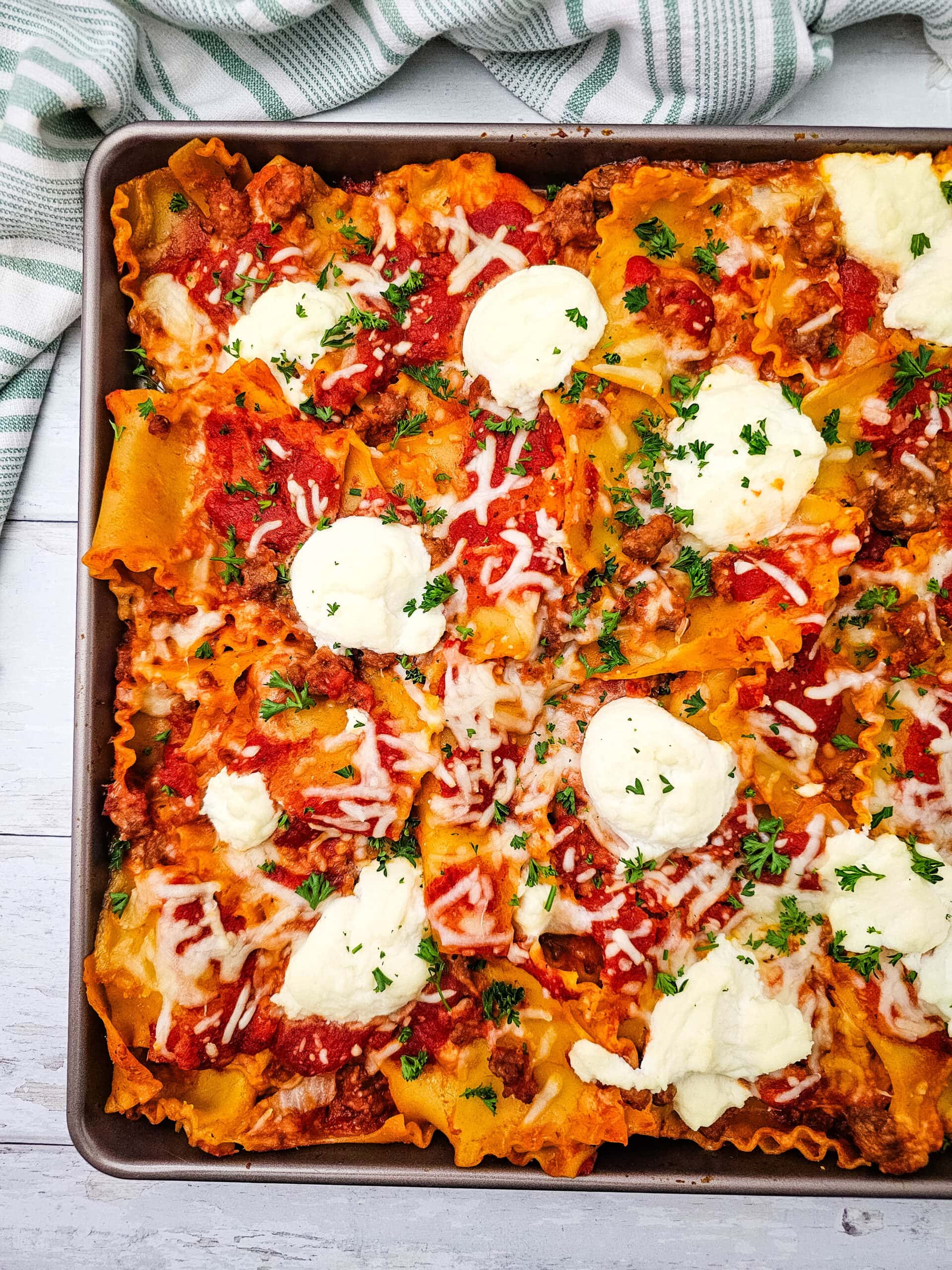 A close-up of a baked lasagna in a pan, topped with tomato sauce, melted cheese, ricotta dollops, ground meat, and fresh parsley, with a striped towel in the background.