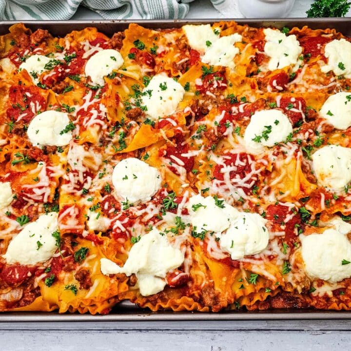 Sheet pan lasagna with wavy pasta, meat sauce, melted cheese, and dollops of ricotta, garnished with chopped parsley and served on a rustic white table.