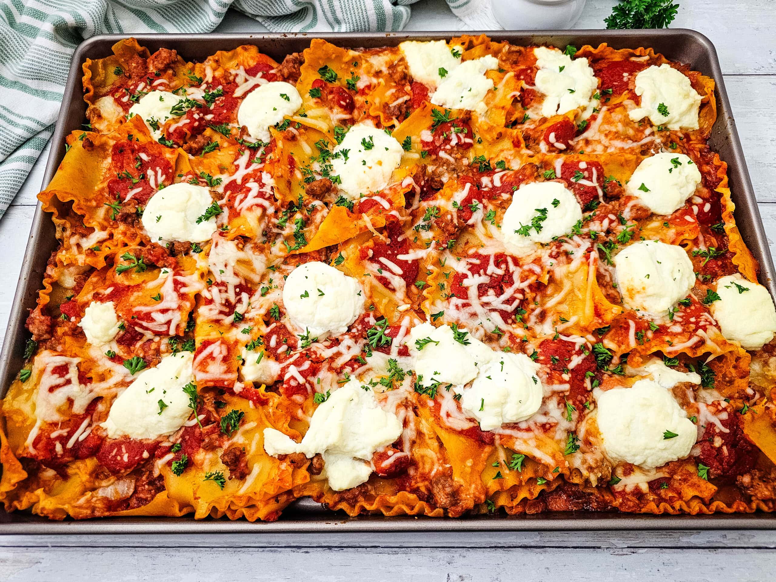 Sheet pan lasagna with wavy pasta, meat sauce, melted cheese, and dollops of ricotta, garnished with chopped parsley and served on a rustic white table.