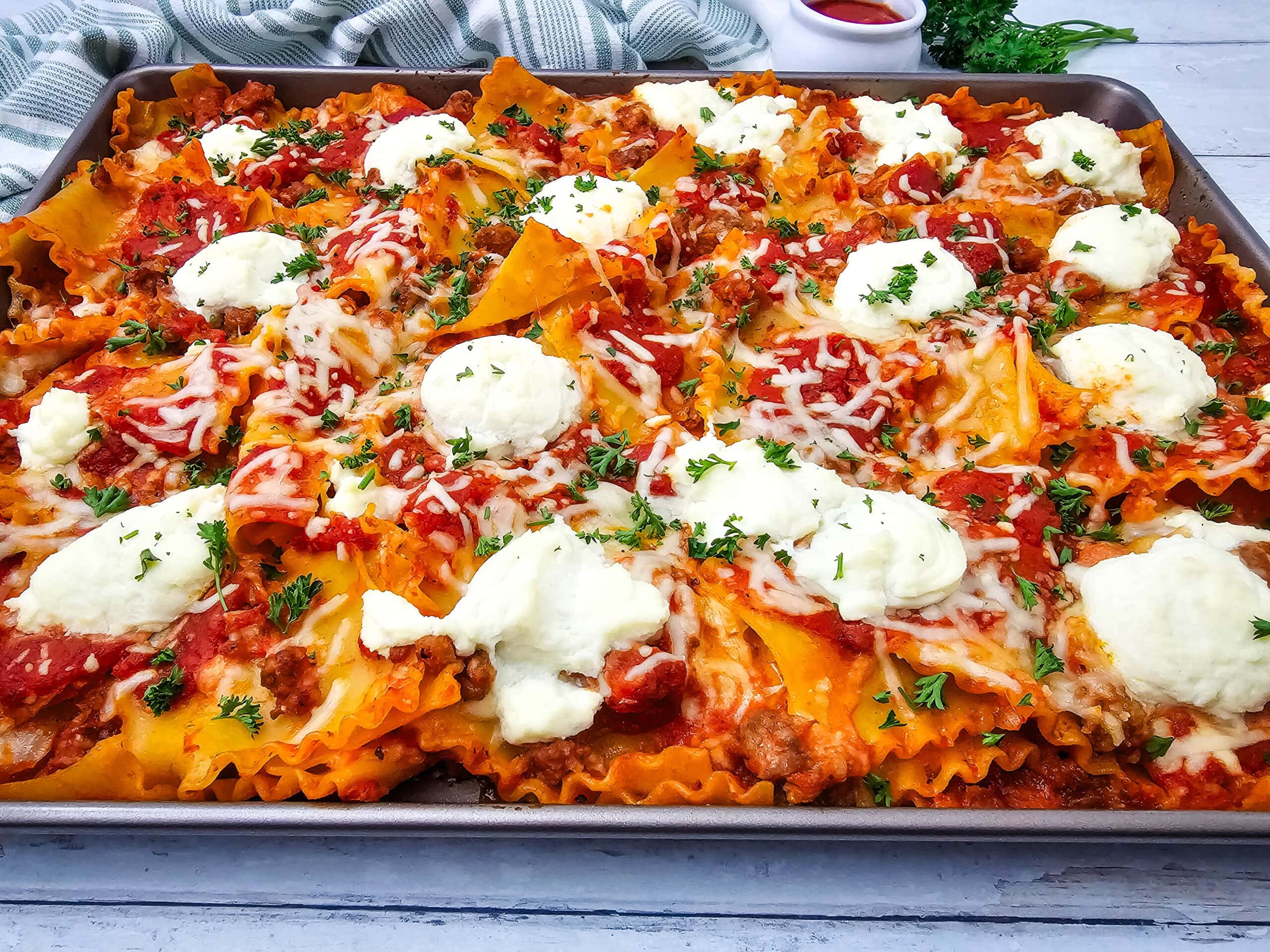 Sheet pan lasagna with wavy noodles, tomato meat sauce, melted mozzarella, and dollops of ricotta, garnished with chopped parsley on a baking tray.