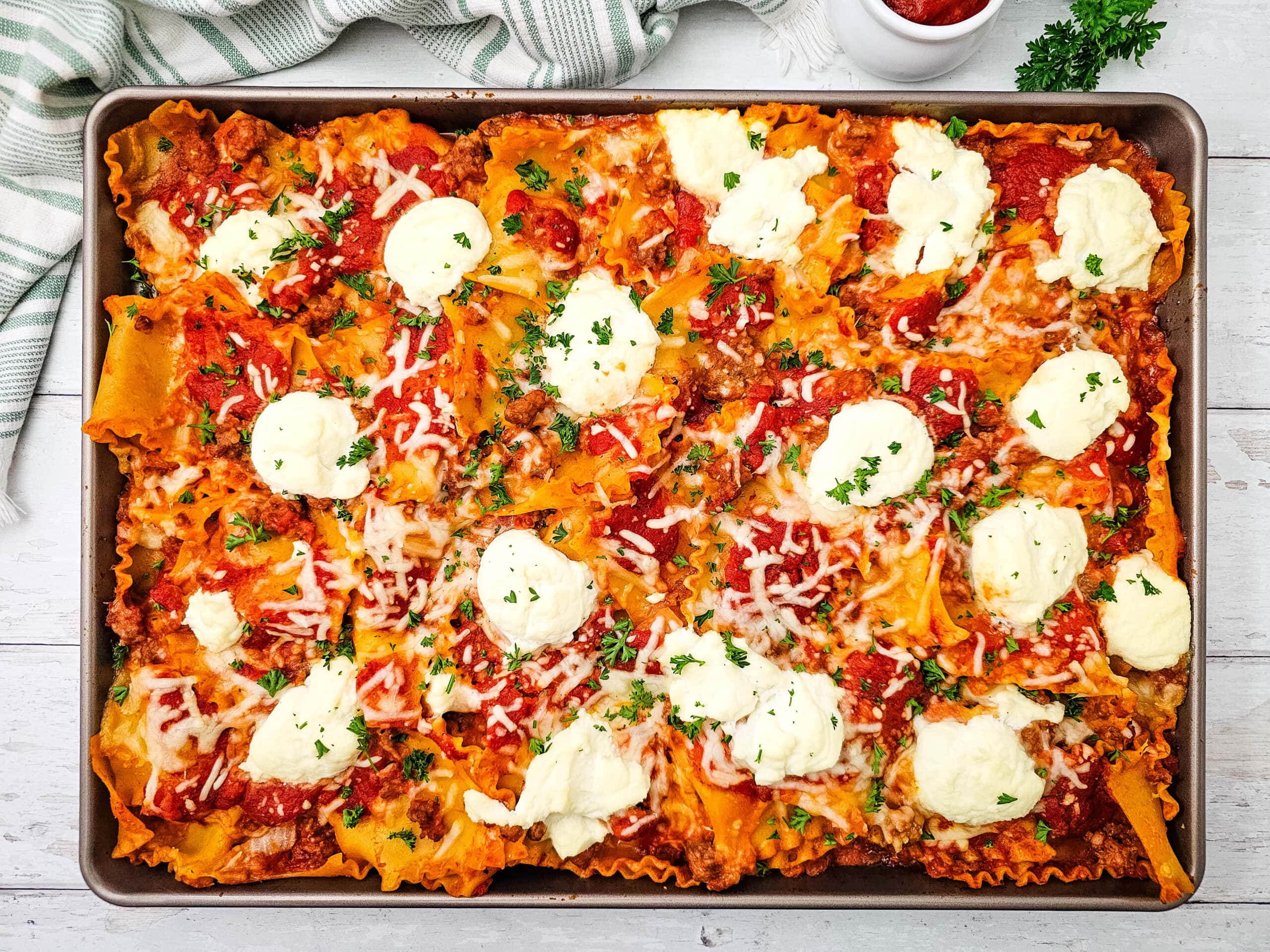 A baking dish filled with baked lasagna topped with tomato sauce, ricotta cheese dollops, melted shredded cheese, and parsley on a white wooden table.