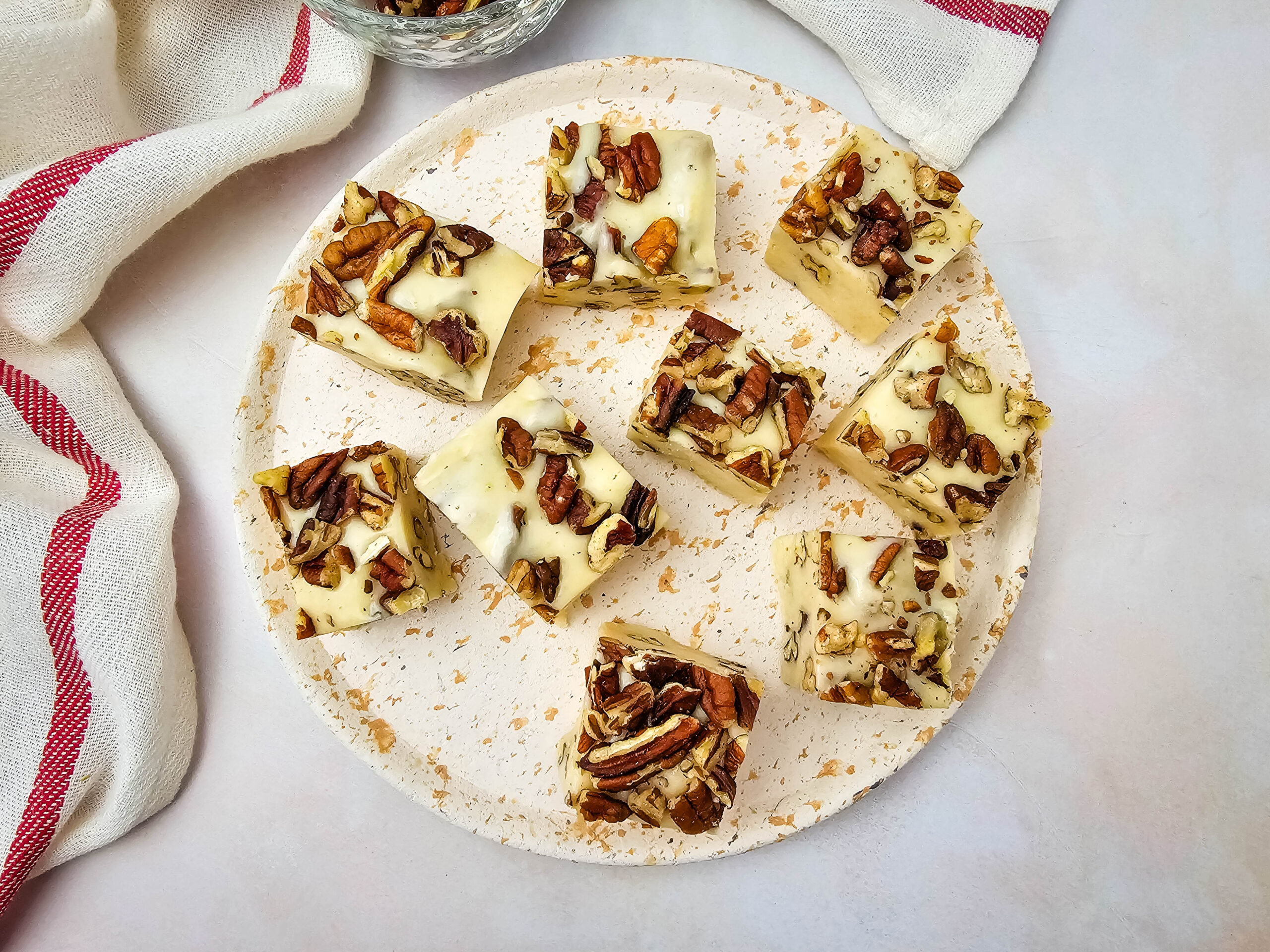 Eight pieces of white chocolate fudge topped with chopped pecans are arranged on a round, white plate. A white towel with red stripes and a bowl with extra pecans are in the background.