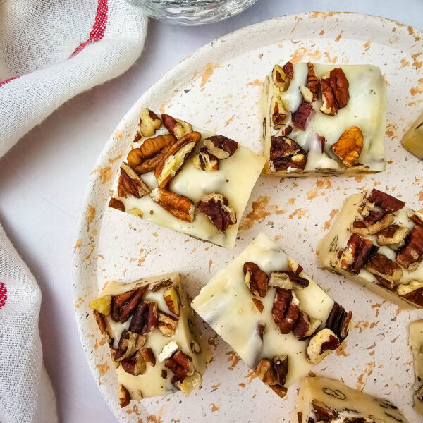 Five white chocolate fudge squares topped with chopped pecans are arranged on a white speckled plate. A small bowl of additional pecans and a white towel with a red stripe are beside the plate.