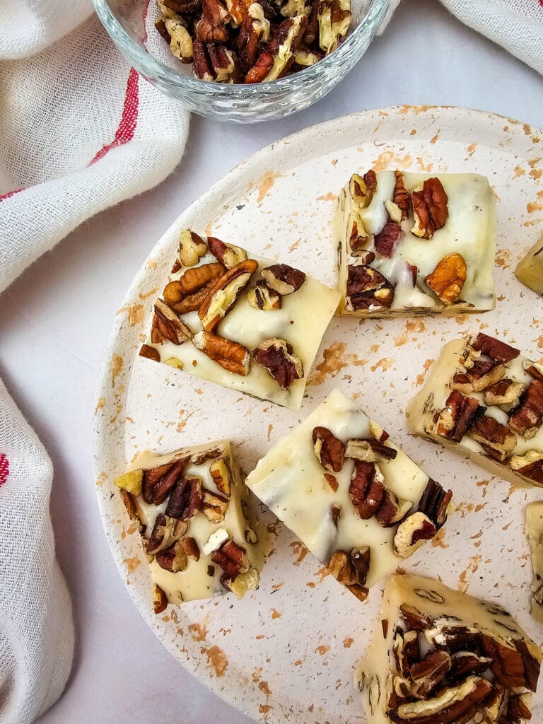 Five white chocolate fudge squares topped with chopped pecans are arranged on a white speckled plate. A small bowl of additional pecans and a white towel with a red stripe are beside the plate.