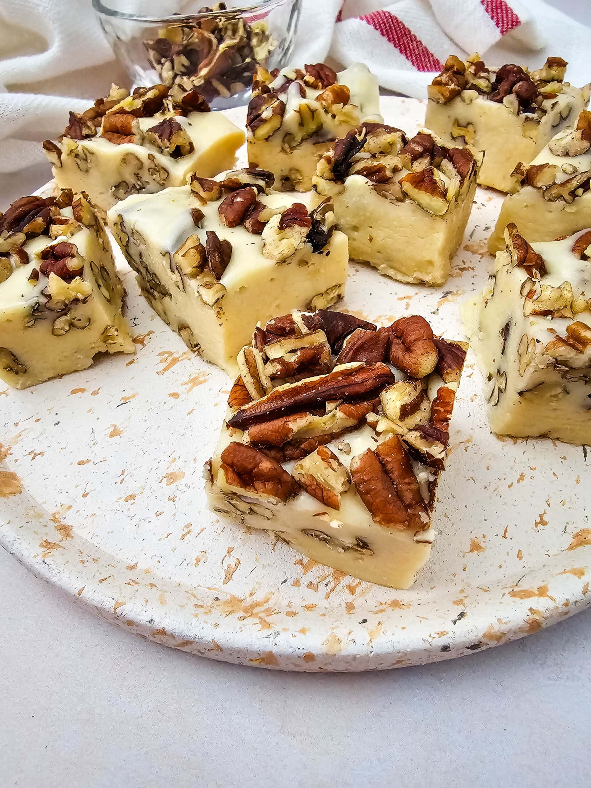 Squares of white fudge with chopped pecans are arranged on a rustic white plate. Some extra pecans are scattered around, and a bowl with more nuts is in the background. A striped cloth is partially visible.