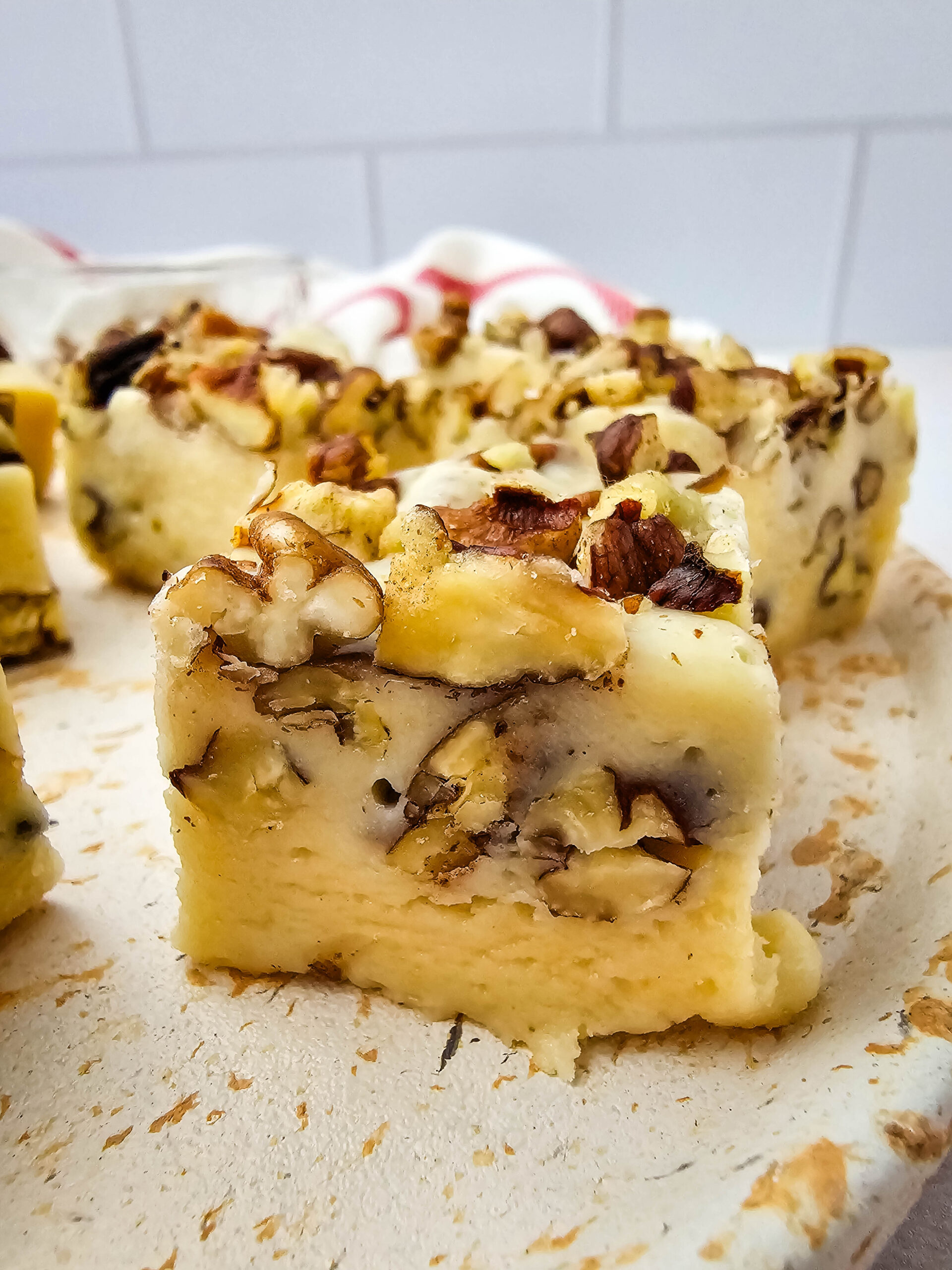 A close-up of a creamy, square cheesecake topped with chopped pecans on a light-colored plate, with more cheesecake pieces and a red-striped towel in the background.