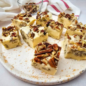Eight pieces of white fudge with chopped pecans sit on a speckled round plate. A small bowl of extra pecans and a red-striped cloth are in the background, all on a light-colored surface.