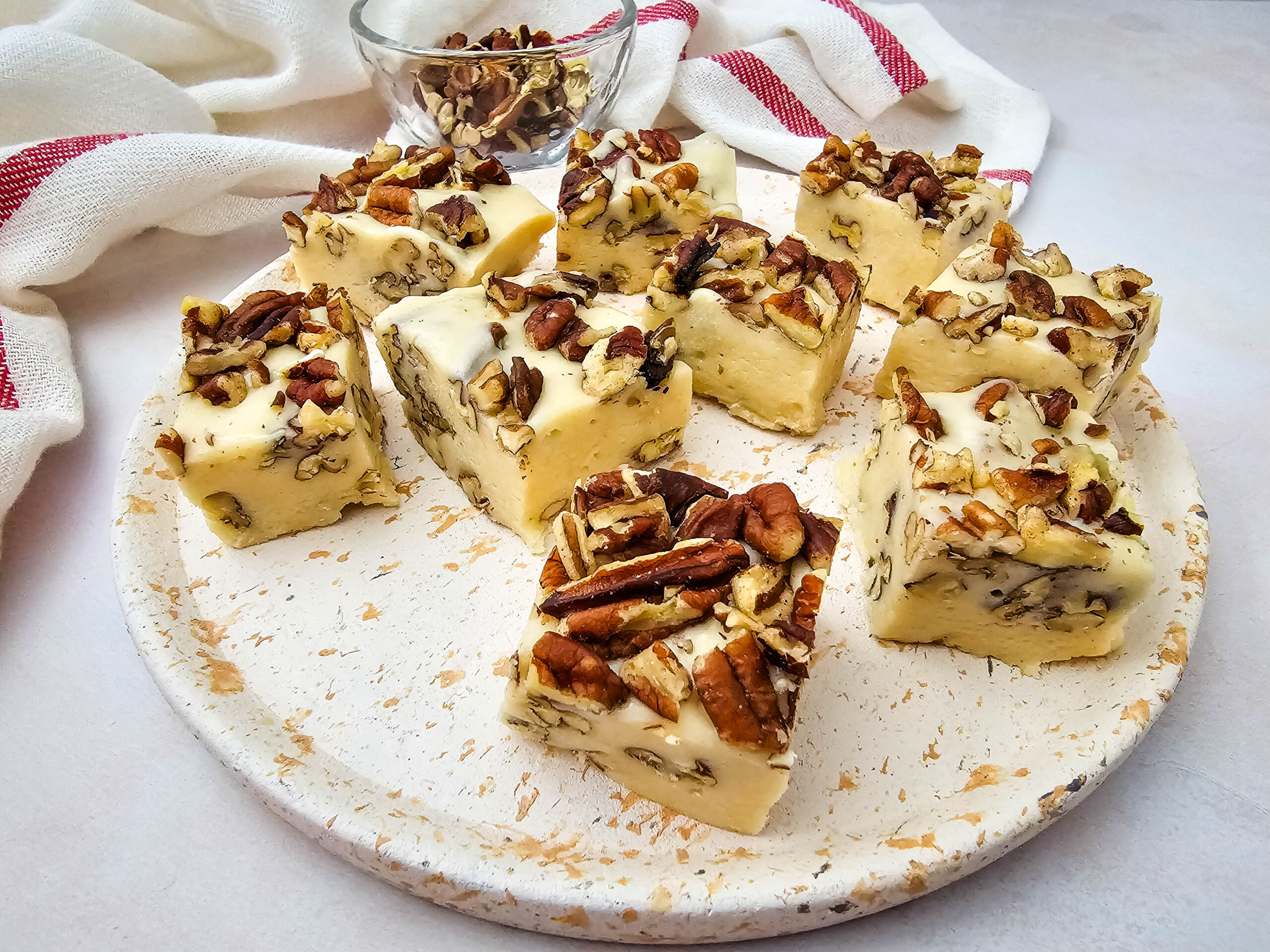Eight pieces of white fudge with chopped pecans sit on a speckled round plate. A small bowl of extra pecans and a red-striped cloth are in the background, all on a light-colored surface.