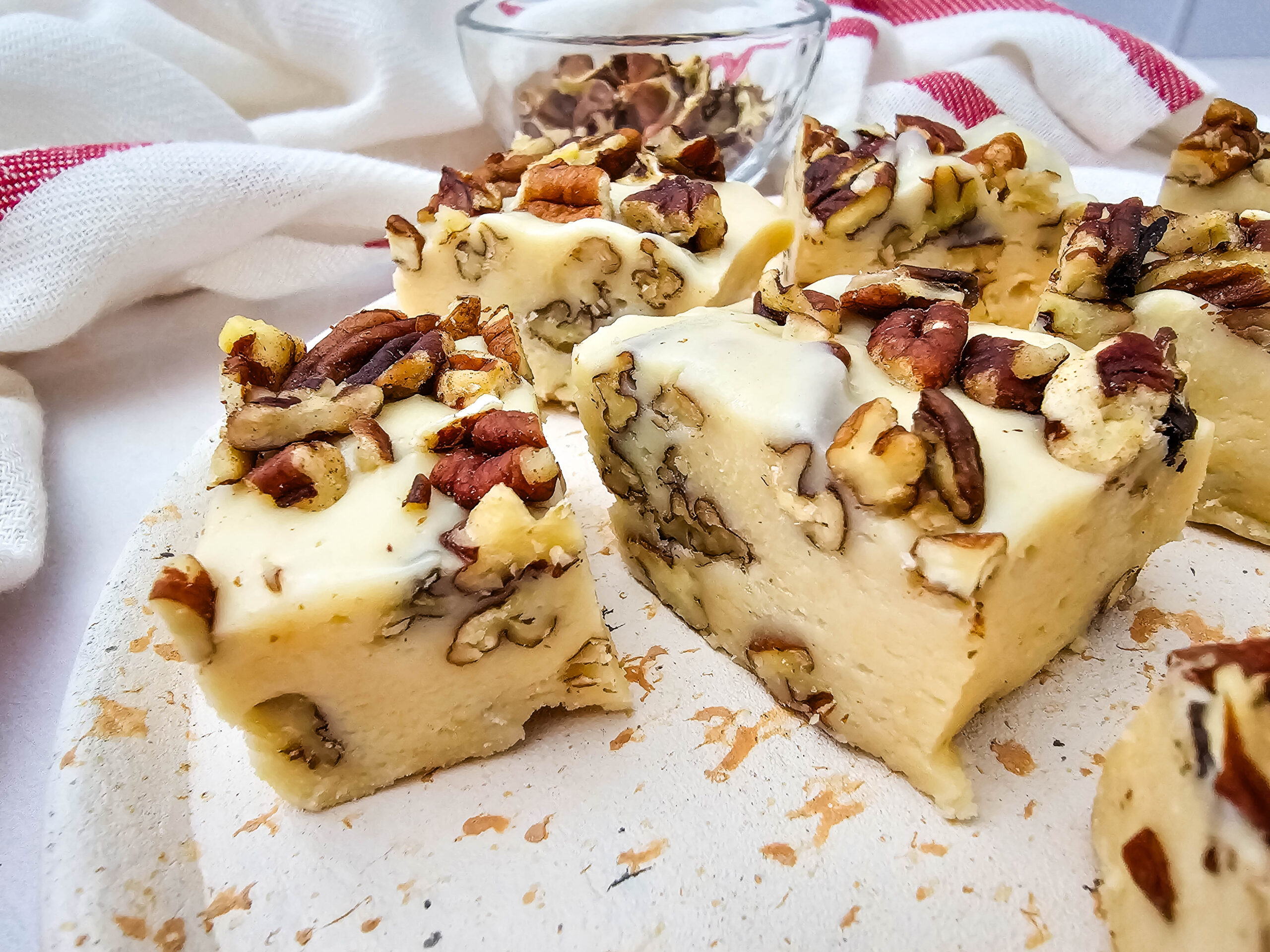 Squares of creamy white fudge topped and filled with chopped pecans are displayed on a light surface, with a white and red cloth and a glass bowl in the background.
