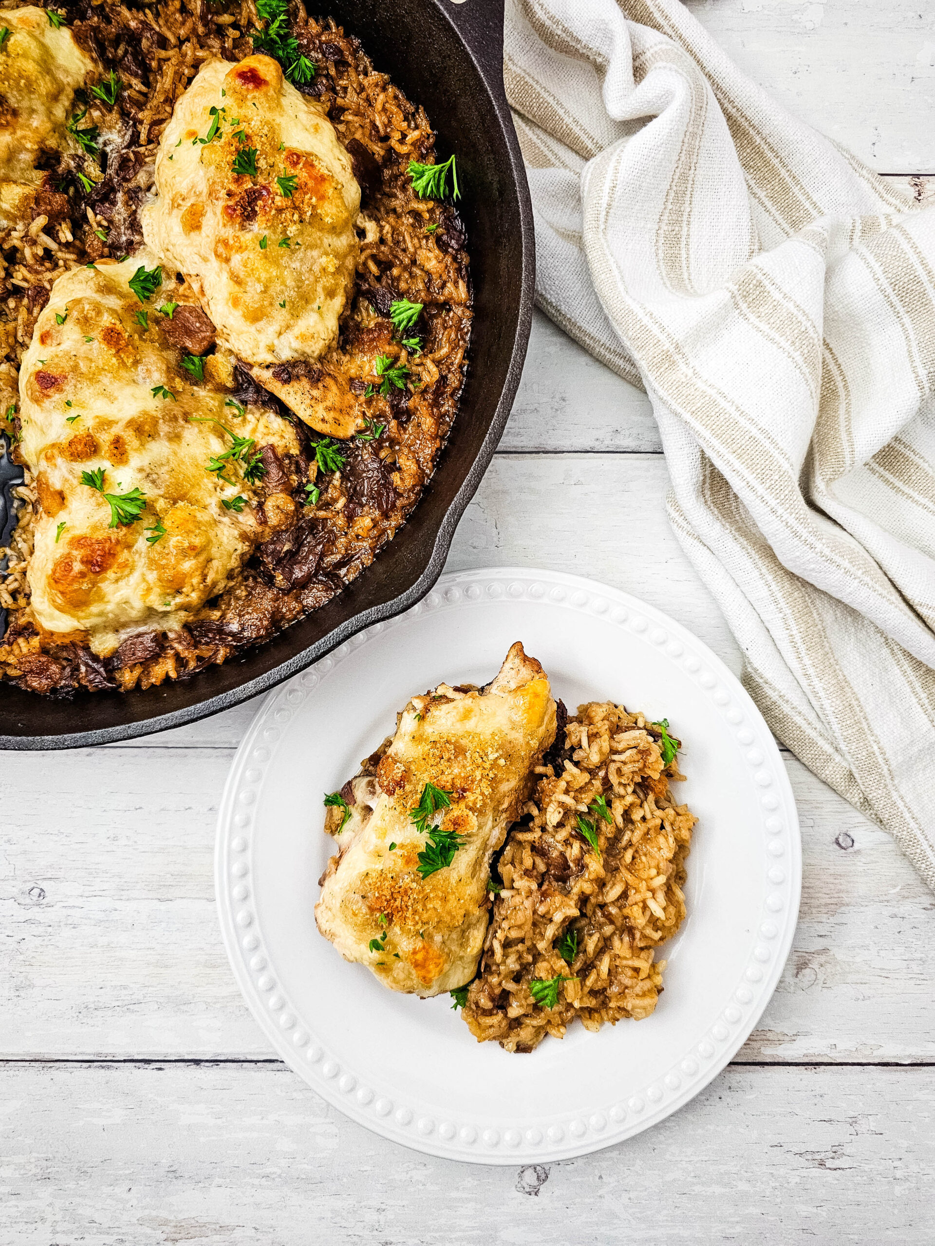 A skillet with baked chicken and rice, garnished with parsley. A plate beside it holds a serving of chicken and rice. A striped cloth lies nearby on a white wooden surface.