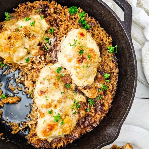 Cast iron skillet with baked chicken breasts on top of seasoned rice, garnished with fresh herbs. A portion is served on a white plate beside the skillet. A beige striped towel is partially visible in the background.