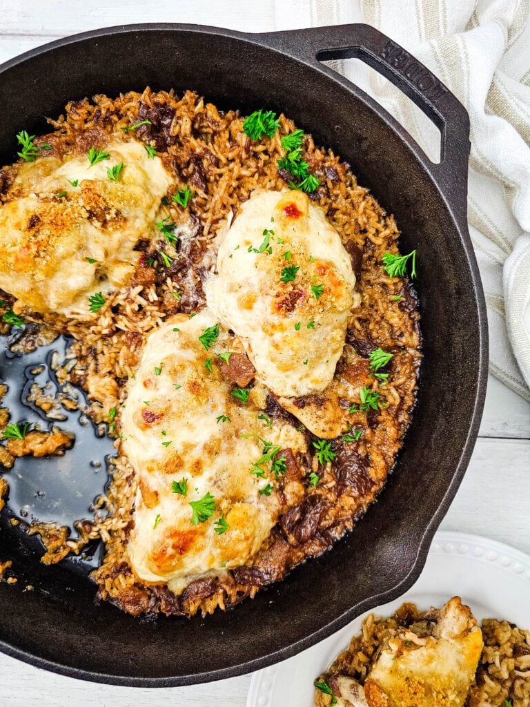 Cast iron skillet with baked chicken breasts on top of seasoned rice, garnished with fresh herbs. A portion is served on a white plate beside the skillet. A beige striped towel is partially visible in the background.