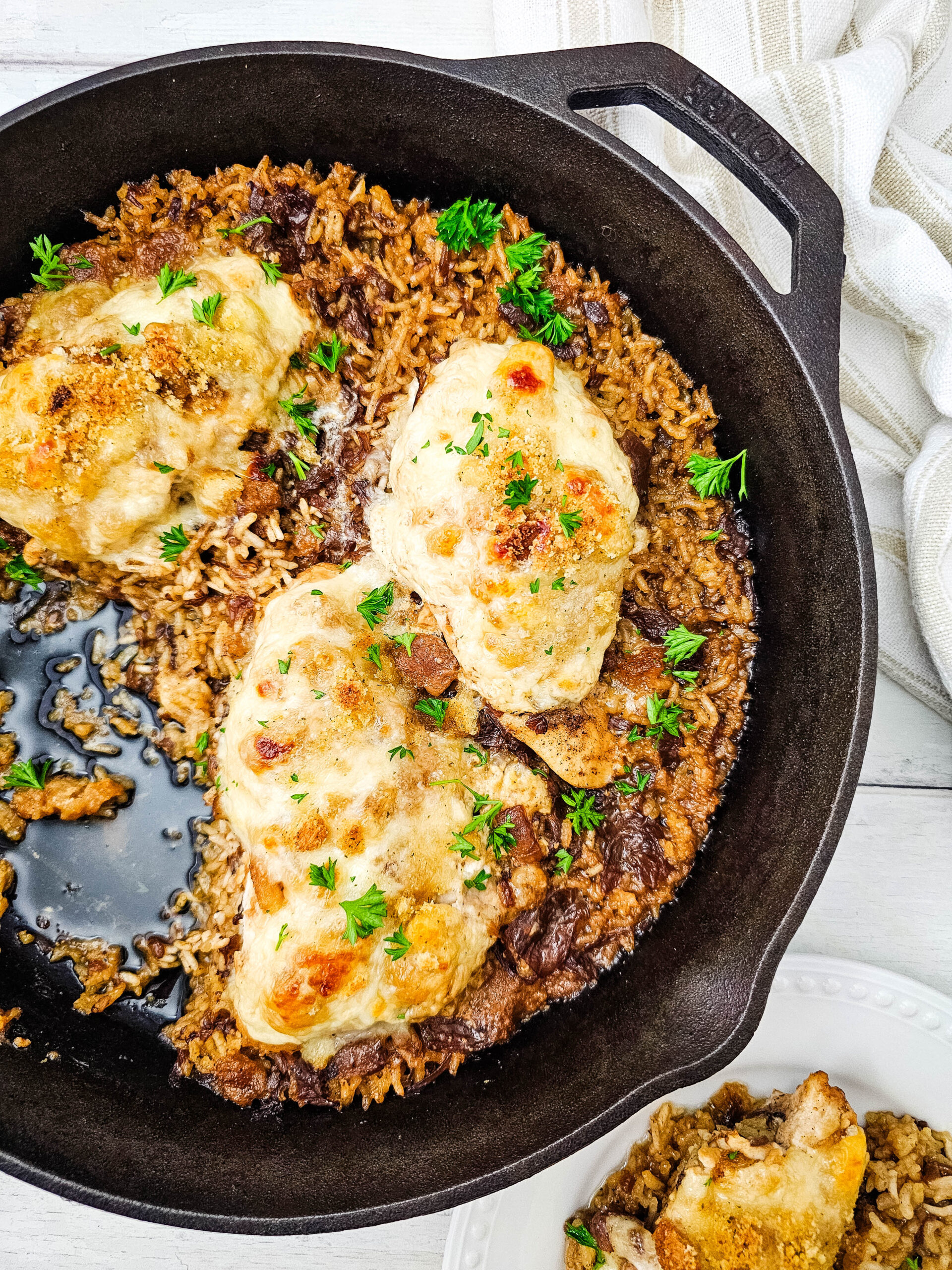 Cast iron skillet with baked chicken breasts on top of seasoned rice, garnished with fresh herbs. A portion is served on a white plate beside the skillet. A beige striped towel is partially visible in the background.
