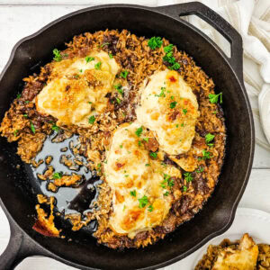 Cast iron skillet with three pieces of baked chicken breast on a bed of seasoned rice, garnished with herbs. A serving of chicken and rice is on a white plate nearby.
