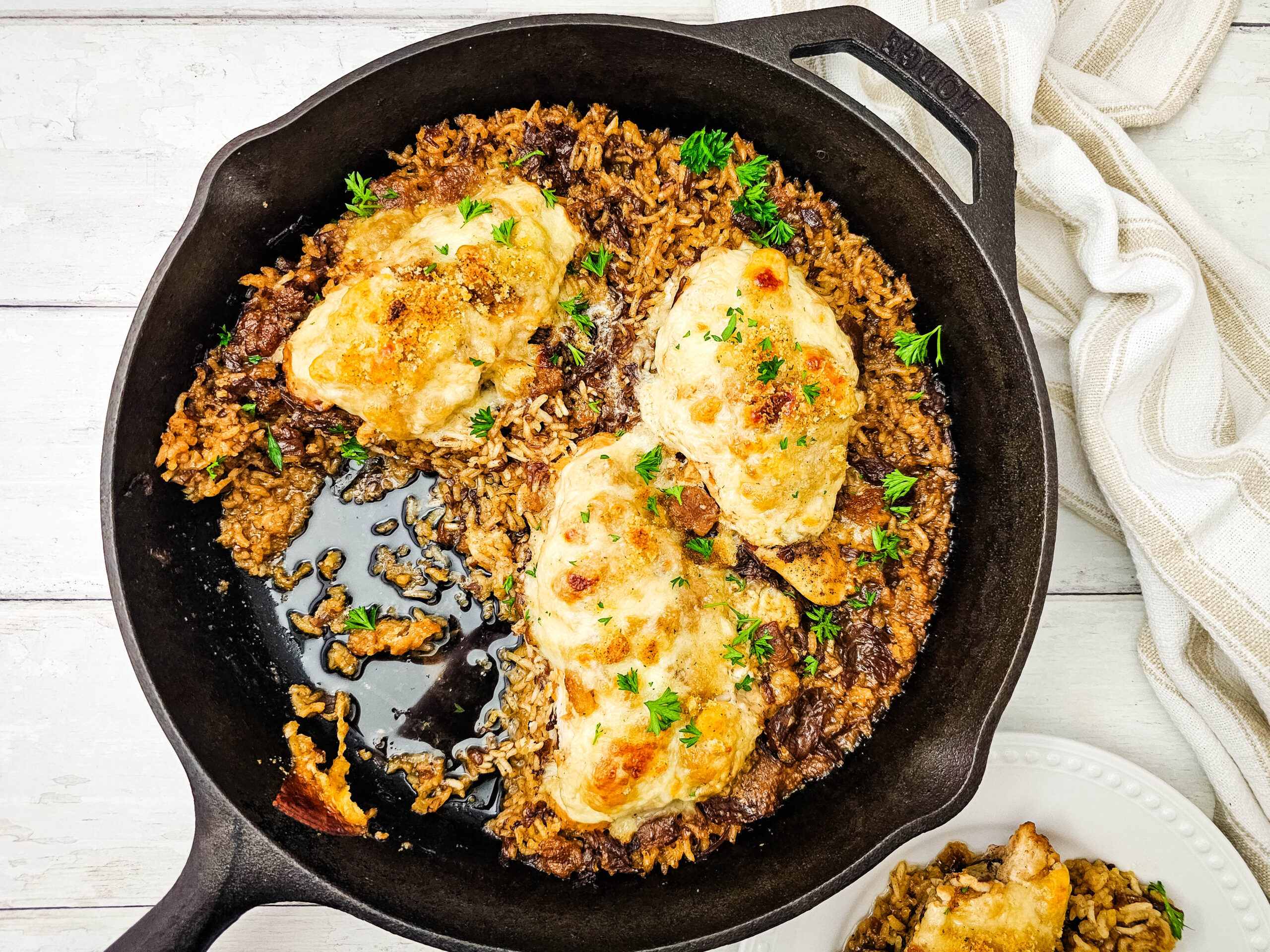 Cast iron skillet with three pieces of baked chicken breast on a bed of seasoned rice, garnished with herbs. A serving of chicken and rice is on a white plate nearby.
