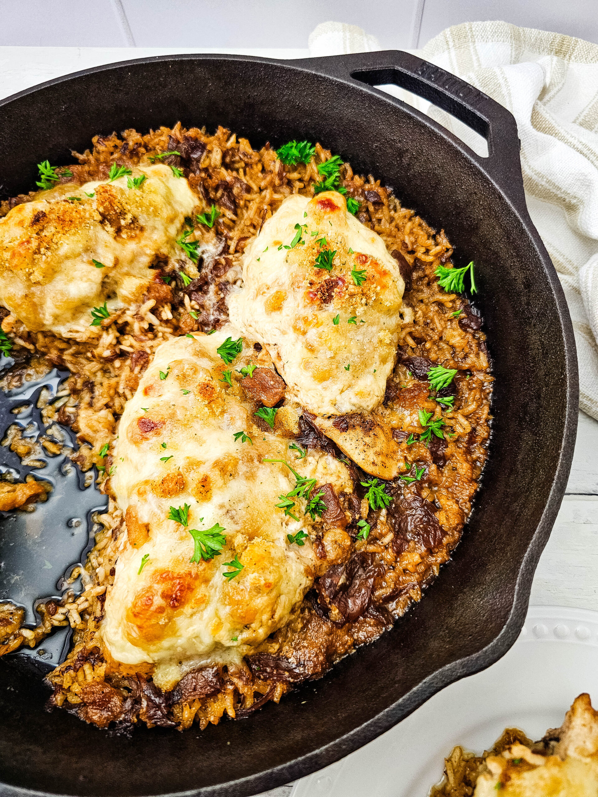 Three baked chicken breasts topping a bed of browned rice in a cast iron skillet, garnished with fresh parsley. A cream-colored towel is nearby.