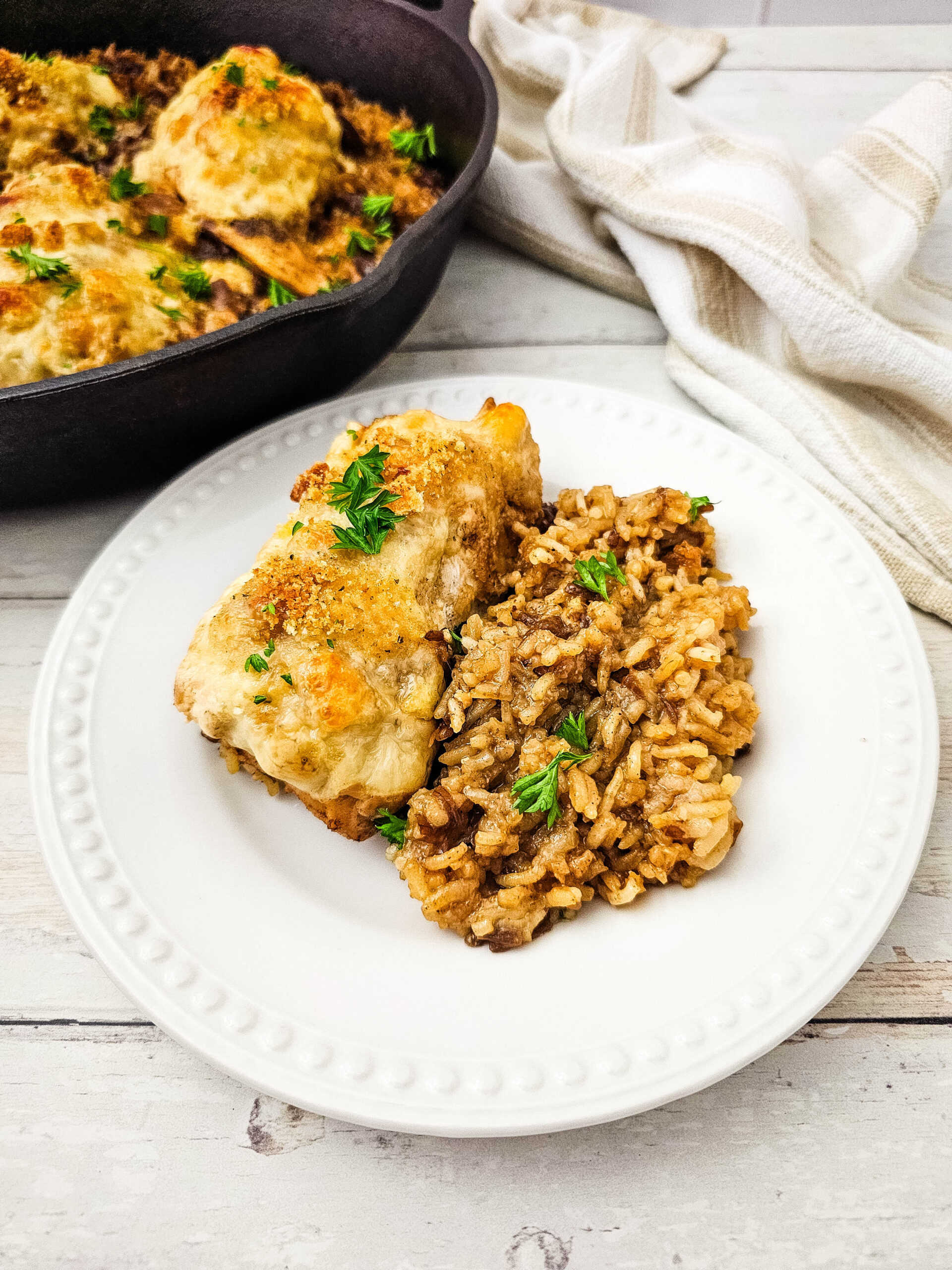 A white plate with baked chicken breast and a serving of seasoned rice, garnished with parsley. A skillet with more chicken and a striped towel are in the background on a light wooden surface.